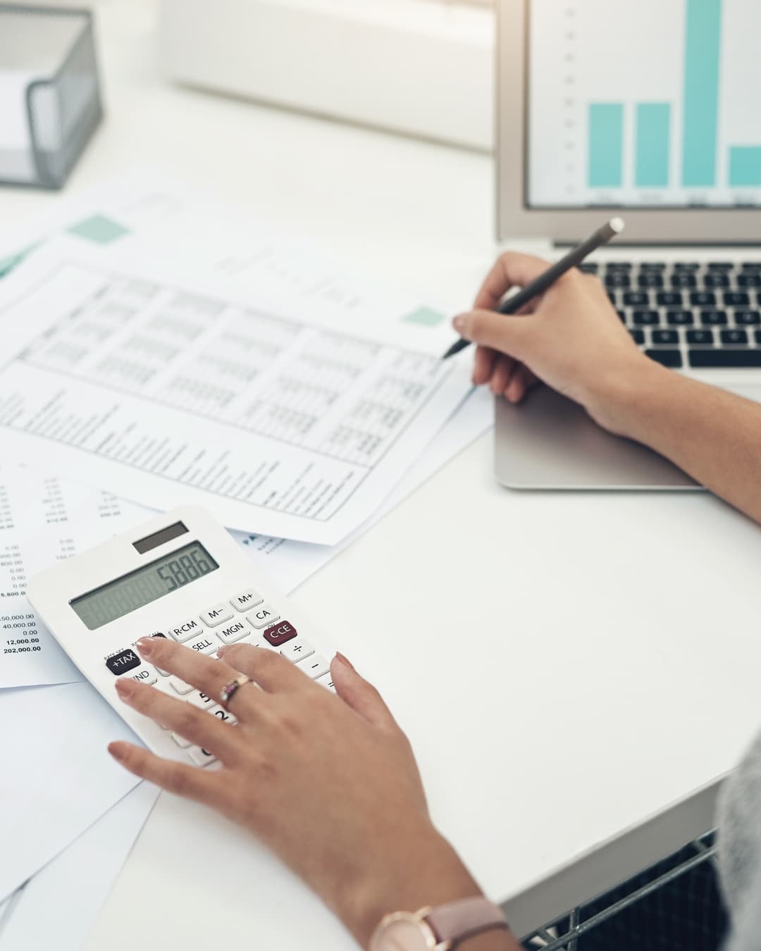 Person using a calculator and writing on documents at a desk with a laptop displaying graphs.